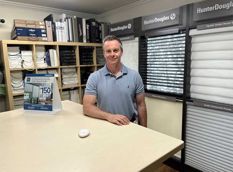 Man in a gray polo standing by a large desk, surrounded by window covering samples